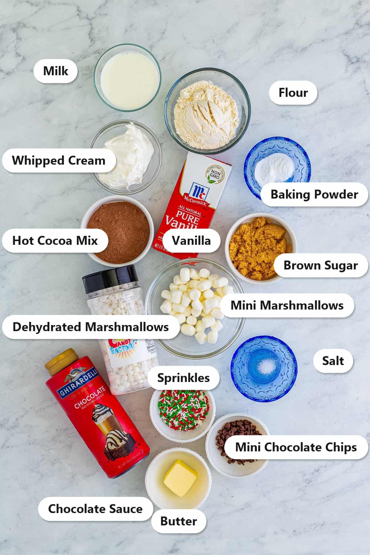 Labeled ingredients for Chocolate Mug Cake including hot cocoa mix, flour, brown sugar, milk, butter, vanilla, mini chocolate chips, marshmallows, and toppings.