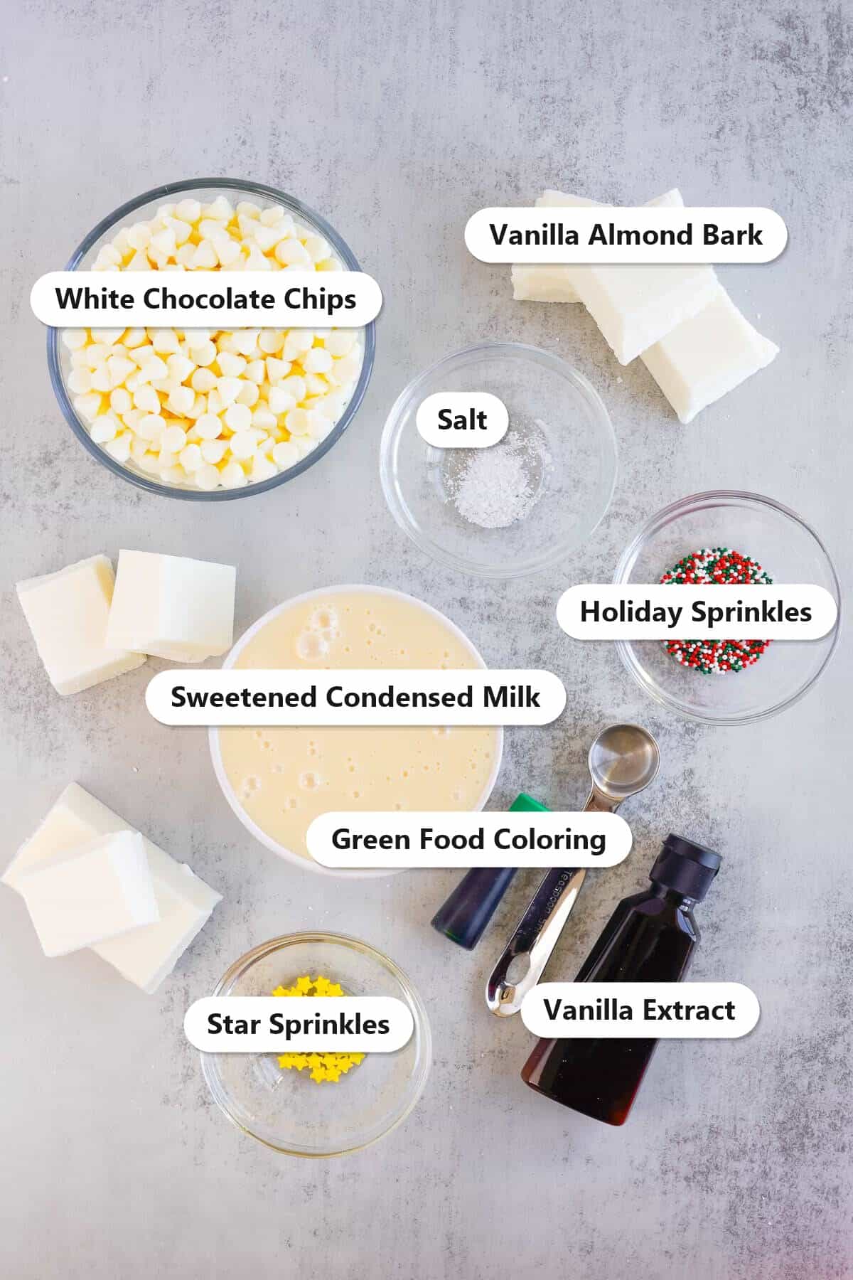 Labeled ingredients for christmas fudge including white chocolate chips, vanilla almond bark, sweetened condensed milk, salt, vanilla extract, green food coloring, and sprinkles.