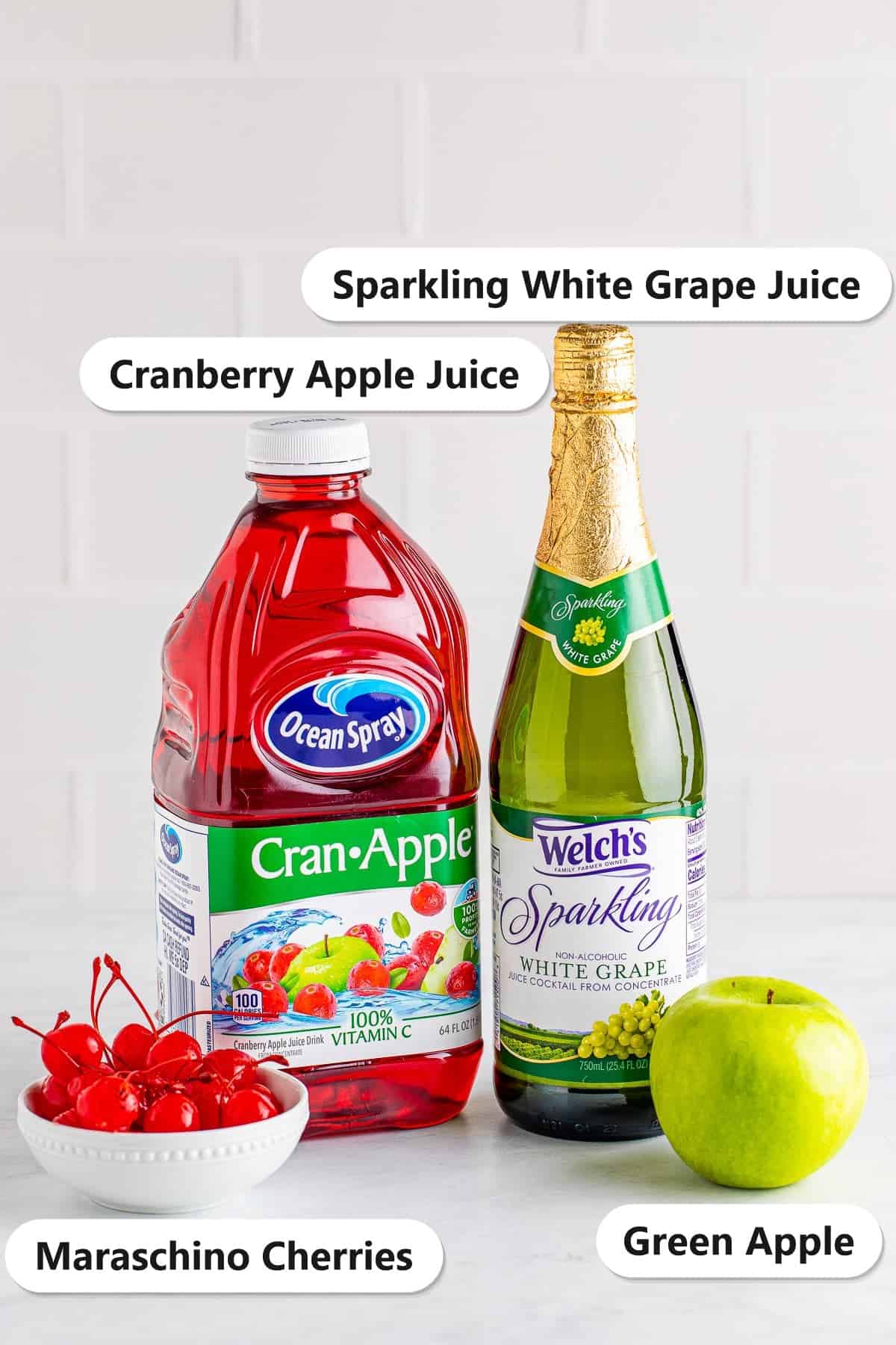 Labeled ingredients for Christmas Mocktails: sparkling white grape juice, cranberry apple juice, maraschino cherries, and a green apple.