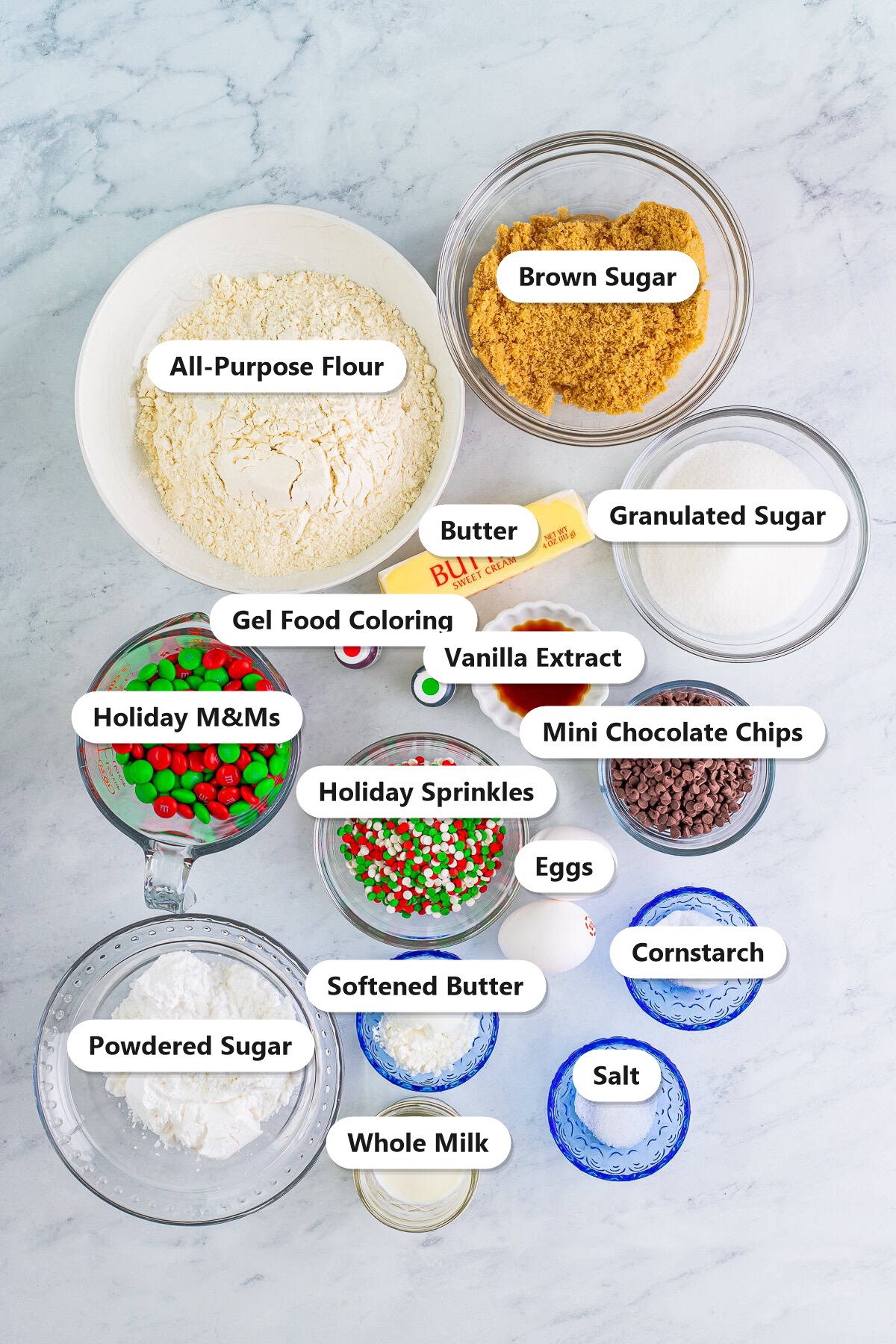 Labeled ingredients for christmas sandwich cookies including flour, butter, sugars, eggs, vanilla, holiday M&Ms, sprinkles, mini chocolate chips, powdered sugar, milk, and food coloring.