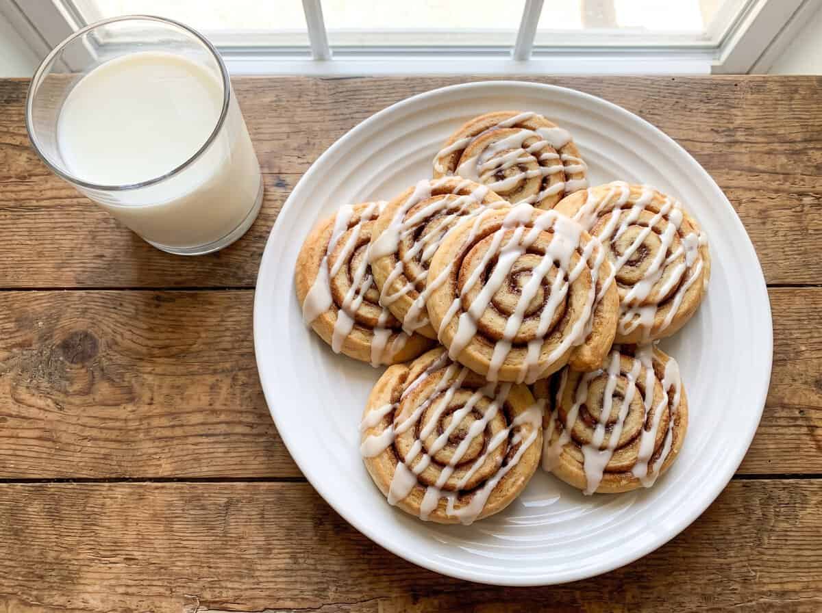 Glazed cinnamon roll cookies arranged on a white plate next to a glass of milk.
