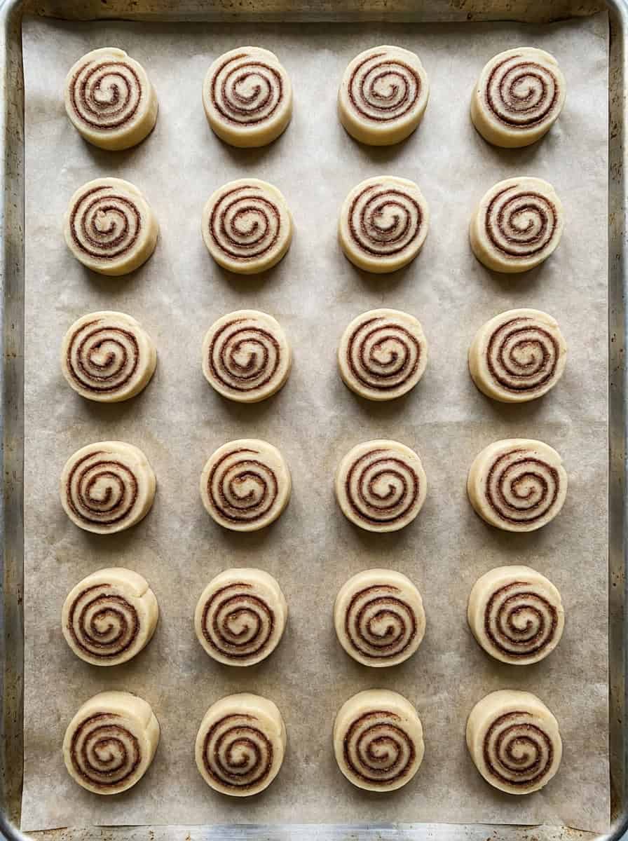 Sliced rounds of cinnamon roll cookie dough with a spiral swirl on a parchment lined baking sheet.