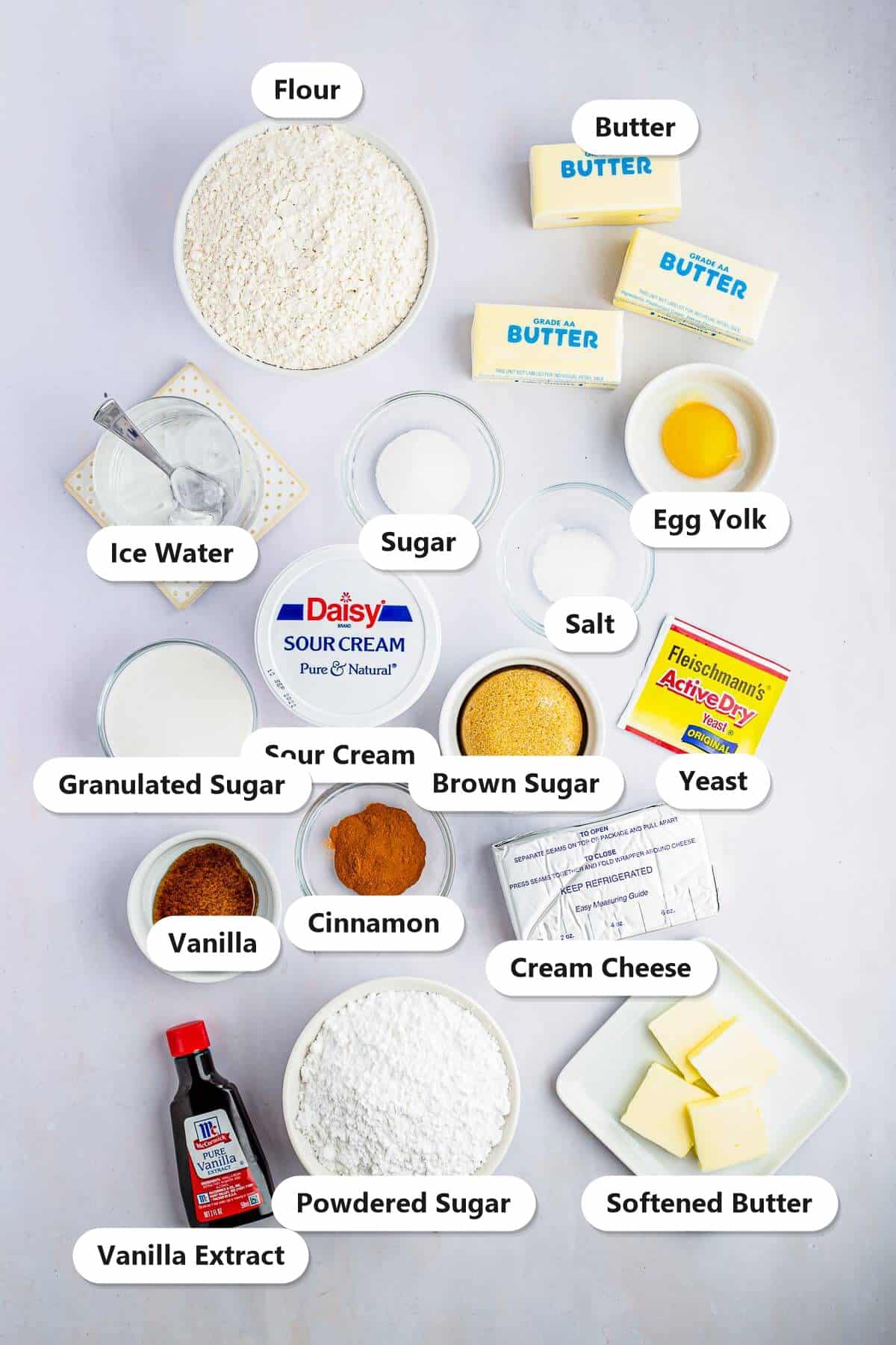 Labeled ingredients to make Cinnamon Rolls: flour, butter, yeast, sugar, sour cream, ice water, egg yolk, brown sugar, cinnamon, vanilla, cream cheese, and powdered sugar.