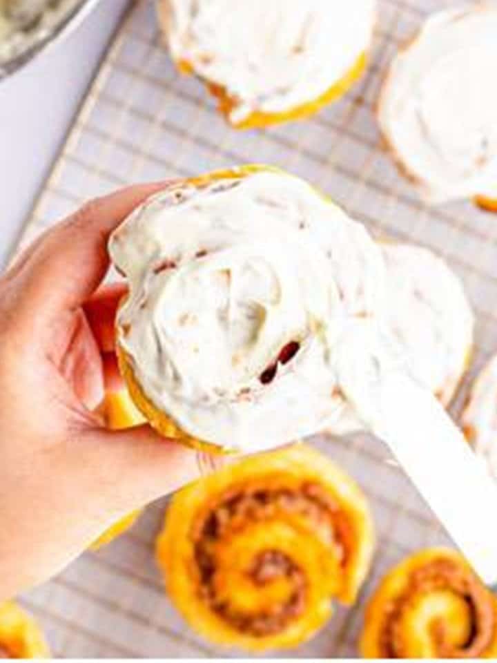 Cream cheese frosting being spread over a baked Cinnamon Roll.