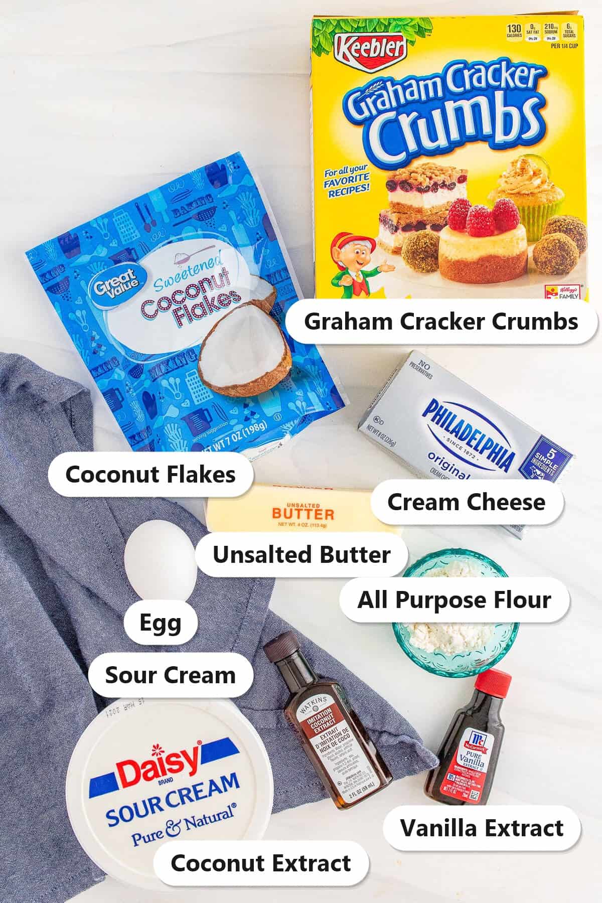 Labeled ingredients for coconut cheesecake bars: graham cracker crumbs, coconut flakes, cream cheese, sour cream, butter, egg, flour, and coconut and vanilla extracts.