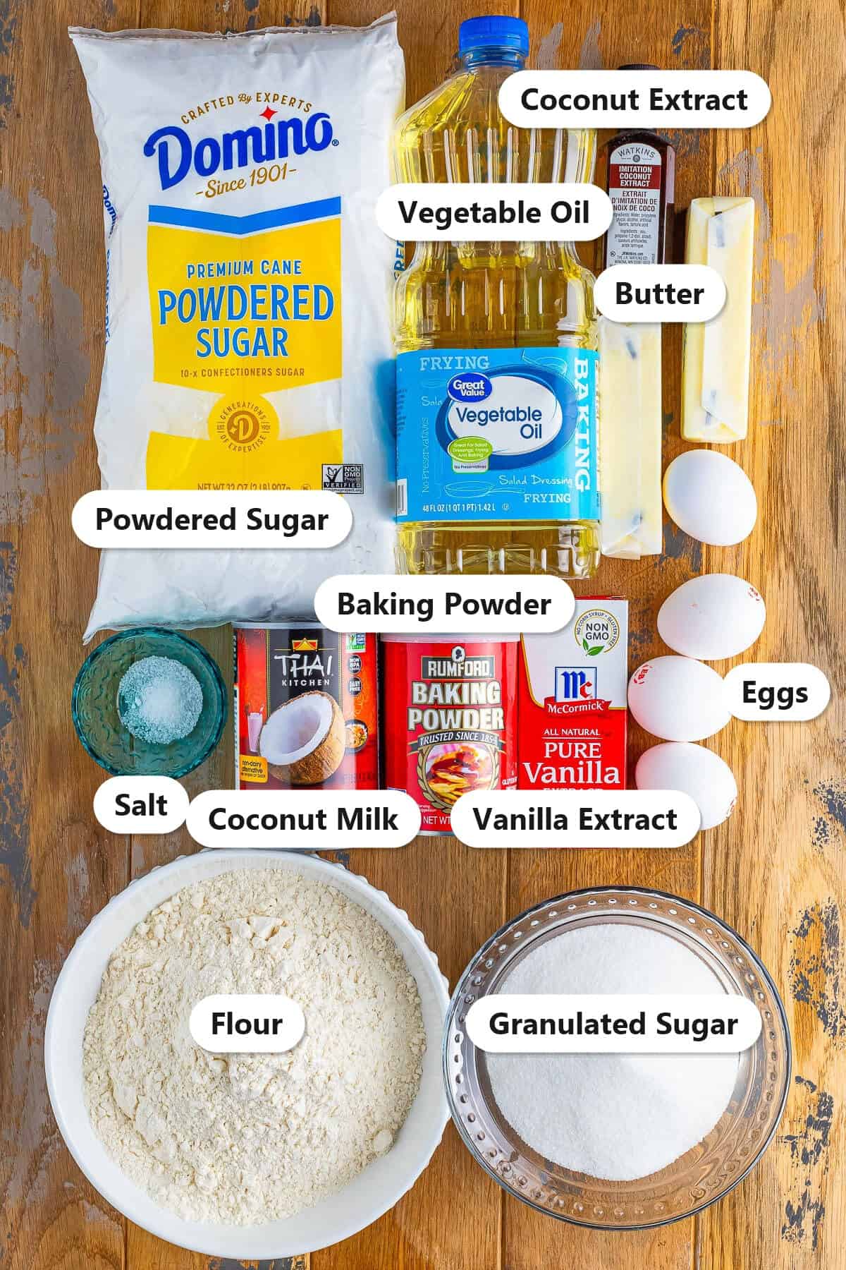 Labeled ingredients for Coconut Cupcakes including flour, granulated sugar, coconut milk, vegetable oil, eggs, coconut extract, vanilla extract, butter, and powdered sugar.