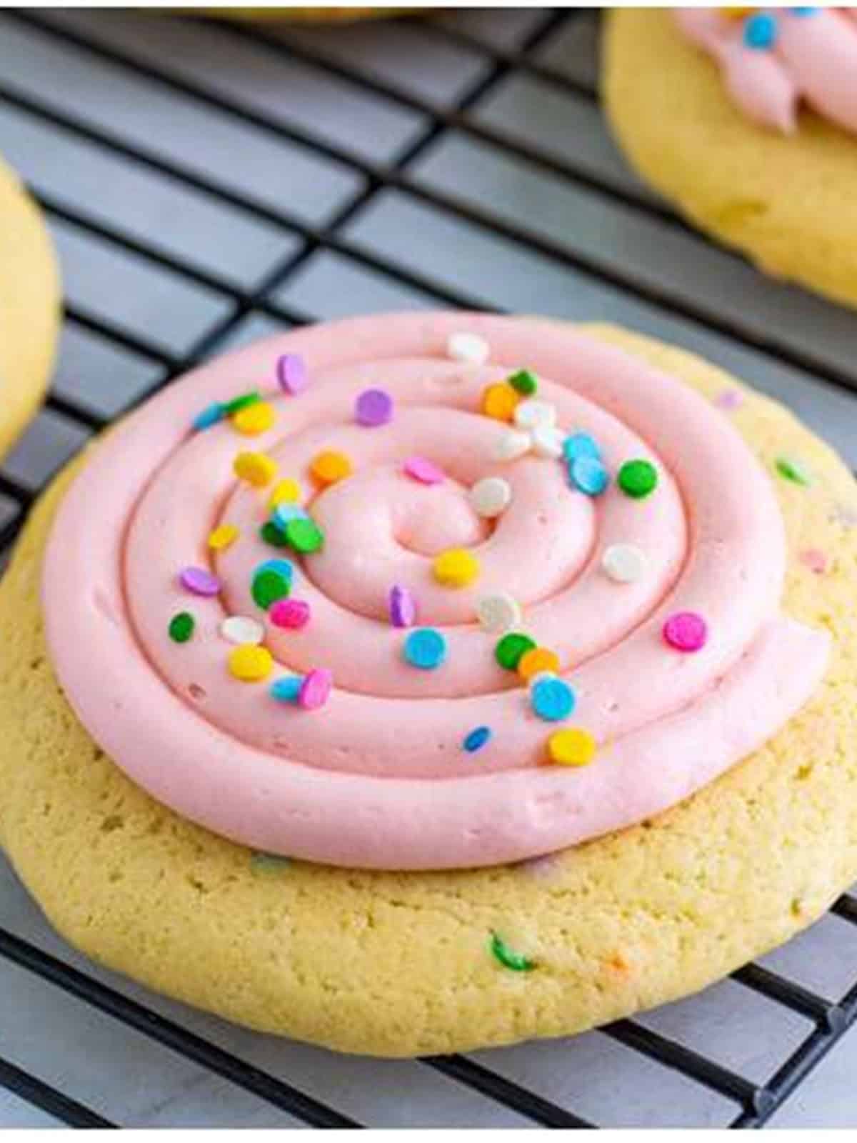 A confetti cake cookie frosted with a swirl of pink frosting and topped with sprinkles on a rack.