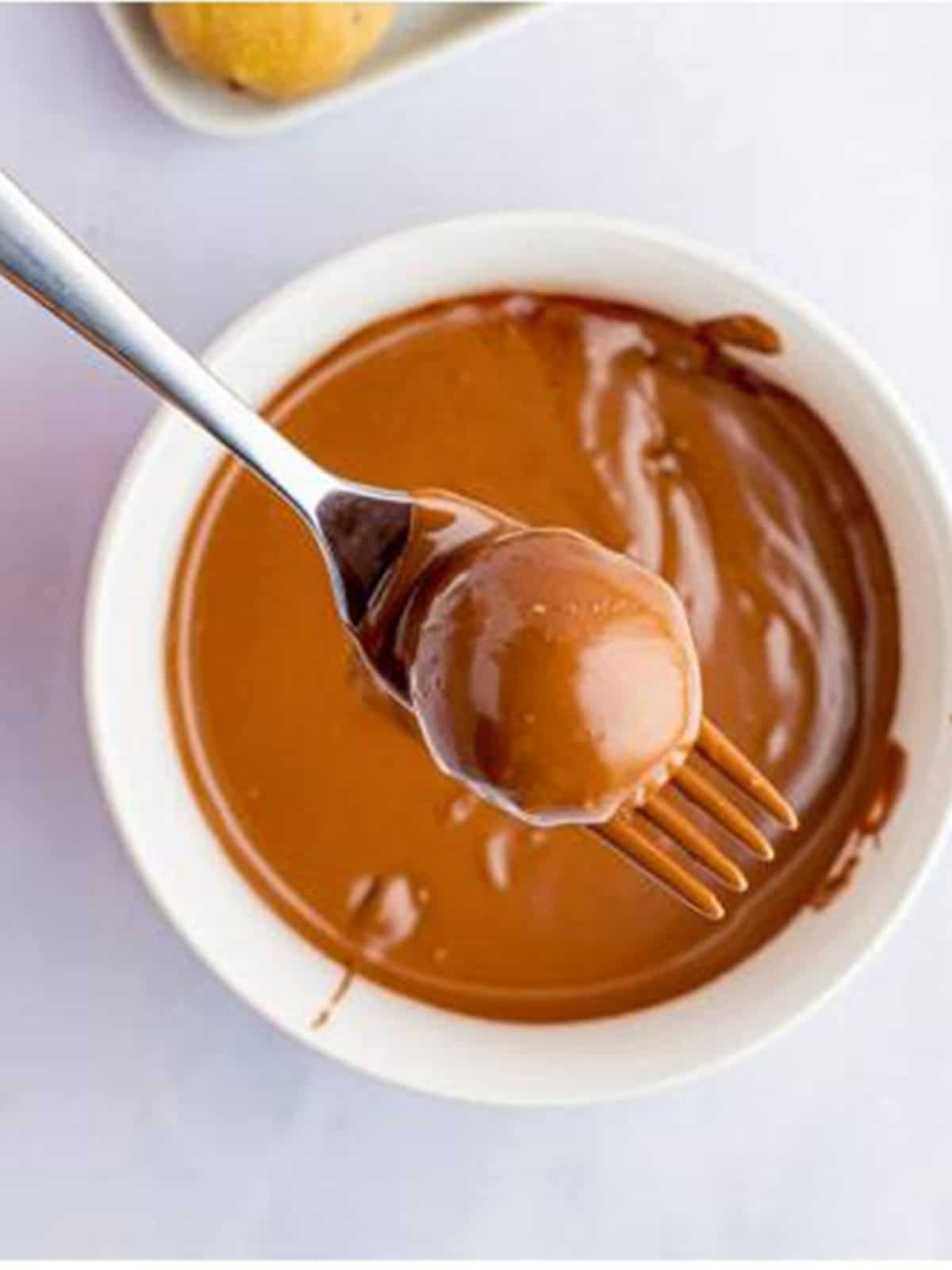 A cookie dough ball being dipped into melted chocolate with a fork.