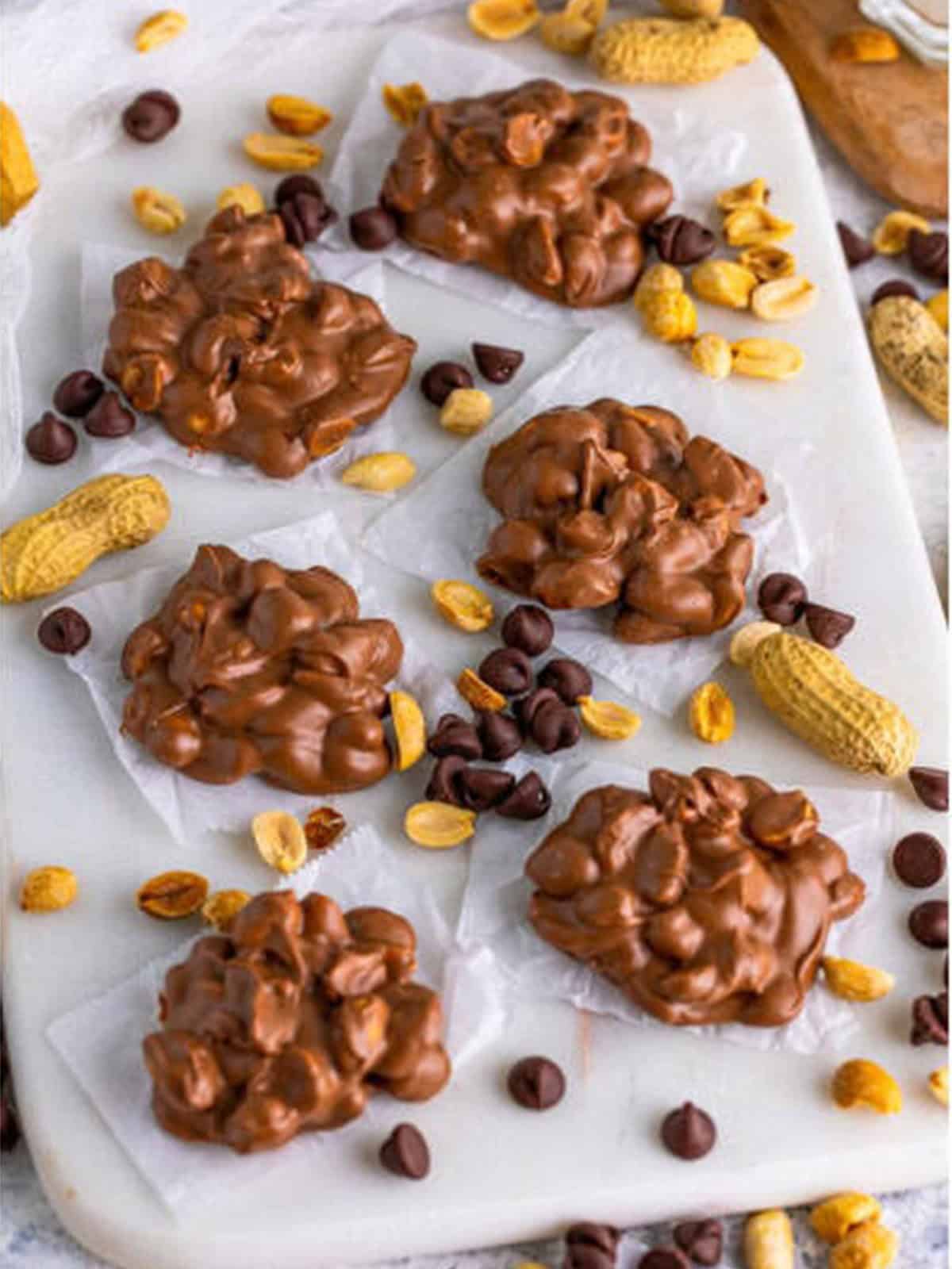 Finished crockpot candy peanut clusters set on a white board with peanuts and chocolate chips