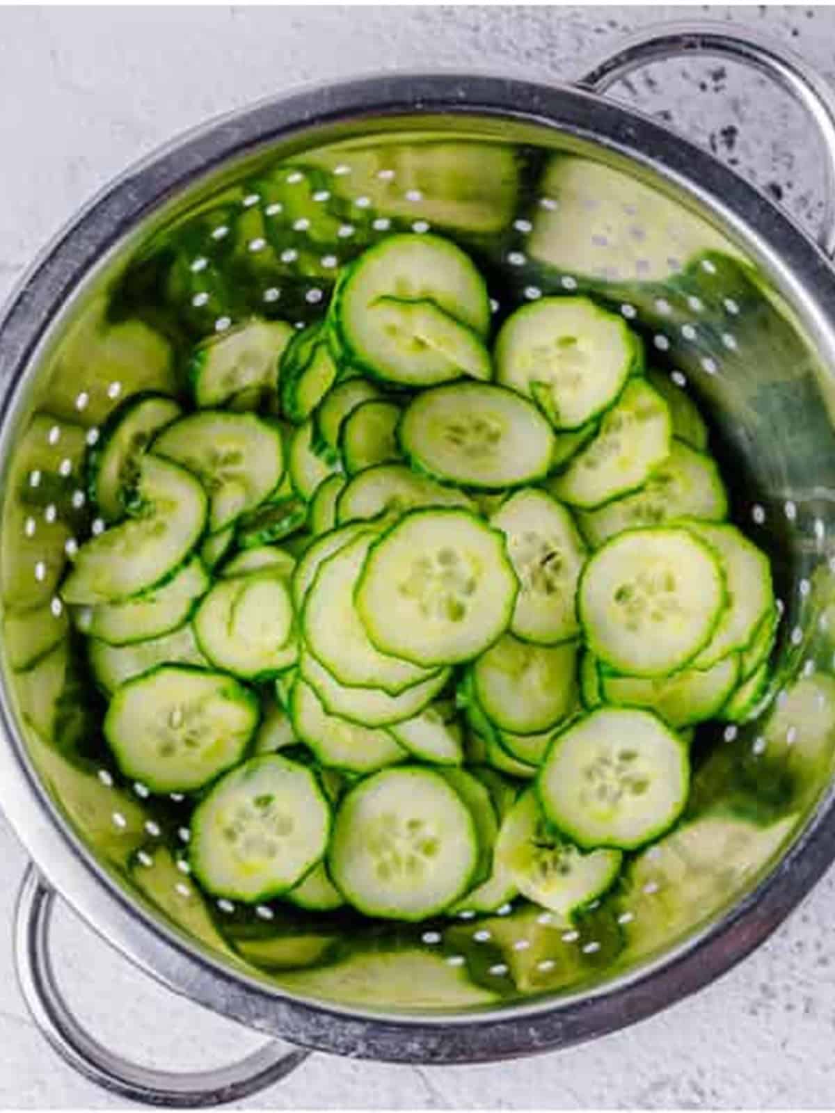 Thinly sliced salted cucumbers draining in a fine mesh strainer for Cucumber Dill Salad.