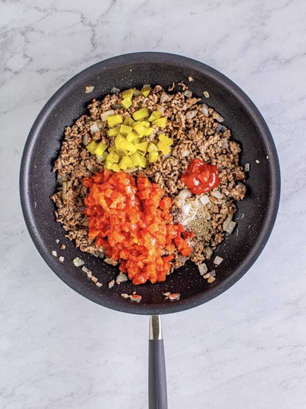 Cooked ground beef and onion in a skillet with diced tomatoes, pickles, and ketchup added on top.