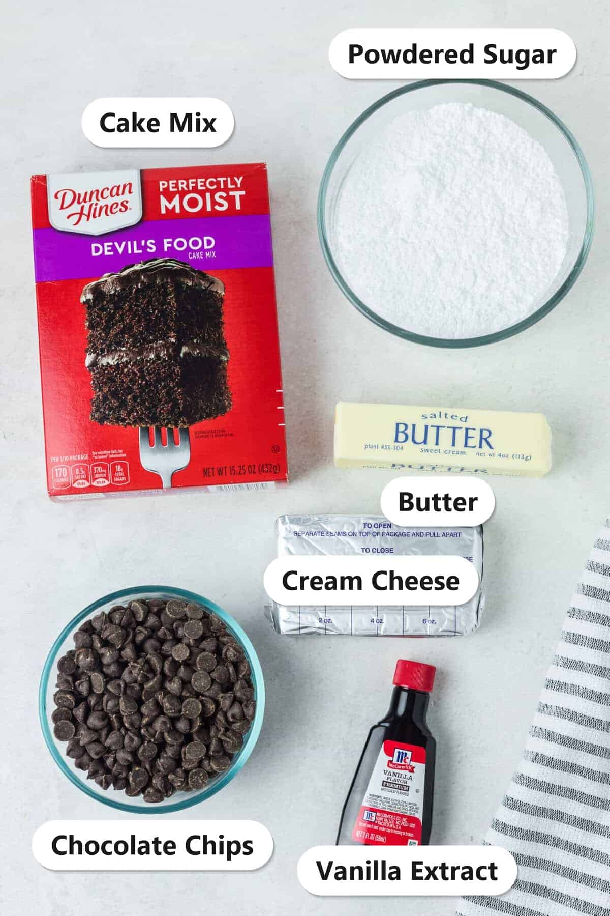 Ingredients to make earthquake cake labeled on a white surface: devils food cake mix, powdered sugar, butter, cream cheese, chocolate chips, and vanilla extract.