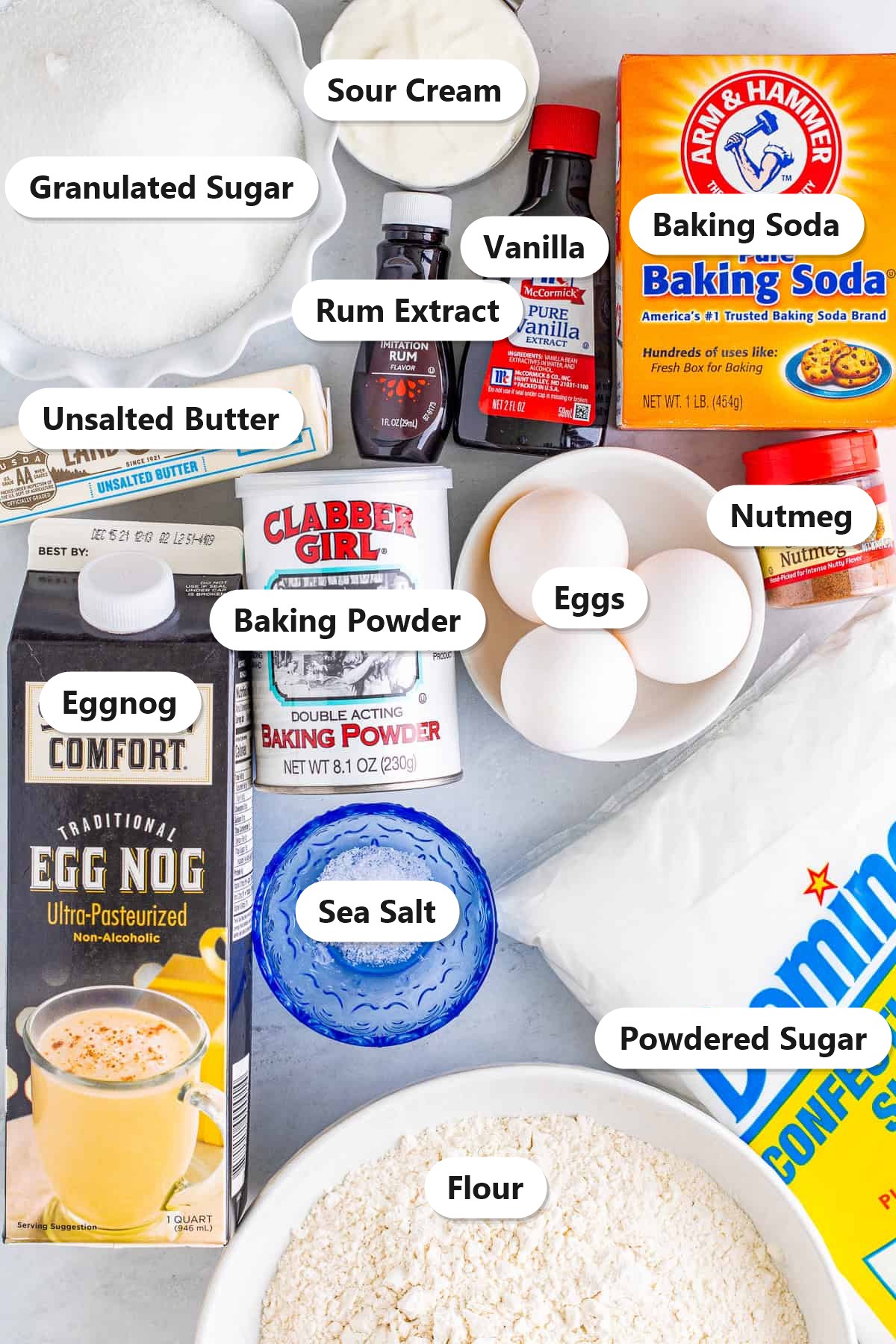 Labeled ingredients for eggnog cake including eggnog, flour, granulated sugar, powdered sugar, butter, eggs, sour cream, vanilla, rum extract, nutmeg, baking soda, baking powder, and sea salt.