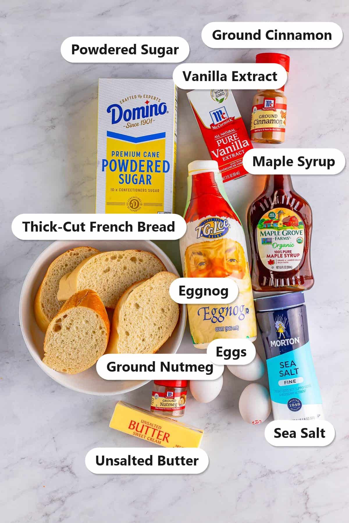 Labeled ingredients for eggnog french toast including eggnog, thick-cut French bread, eggs, cinnamon, nutmeg, vanilla extract, butter, maple syrup, and powdered sugar.