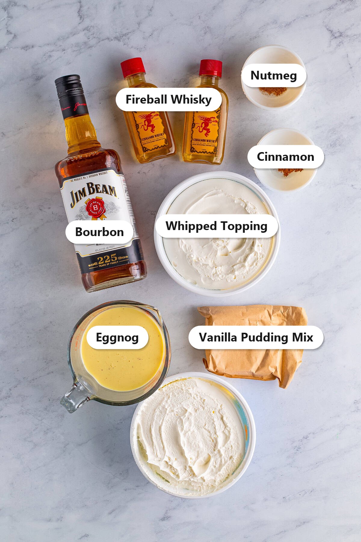 Labeled ingredients for eggnog pudding shots including vanilla pudding mix, eggnog, bourbon, Fireball whisky, cinnamon, nutmeg, and whipped topping.