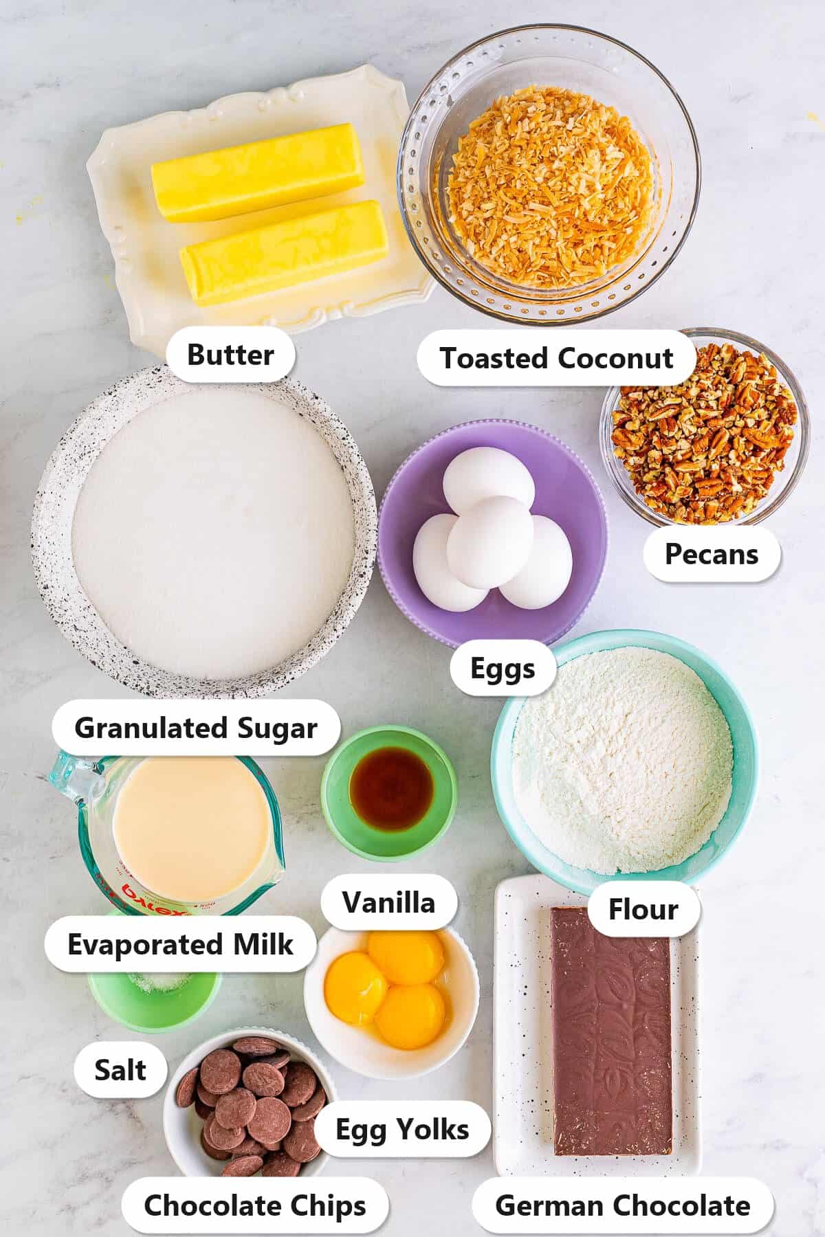 Ingredients to make German chocolate brownies: butter, German chocolate, sugar, eggs, flour, vanilla, salt, evaporated milk, egg yolks, toasted coconut, pecans, and chocolate chips.