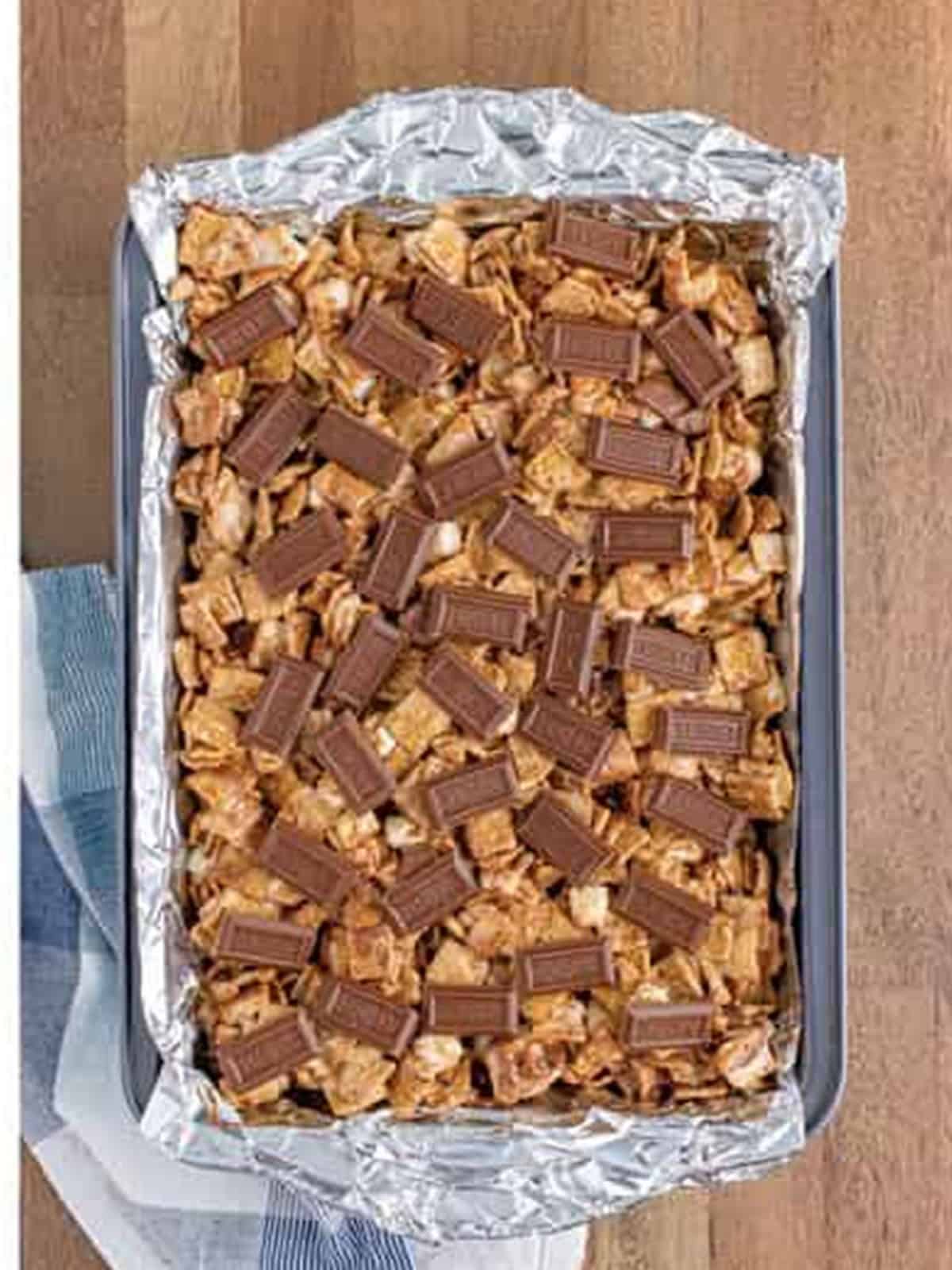 Golden grahams smores bars mixture pressed into a foil lined baking dish and topped with chocolate pieces.