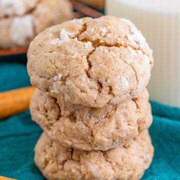 A stack of spiced gooey butter cookies coated in powdered sugar with cinnamon sticks and a glass of milk.