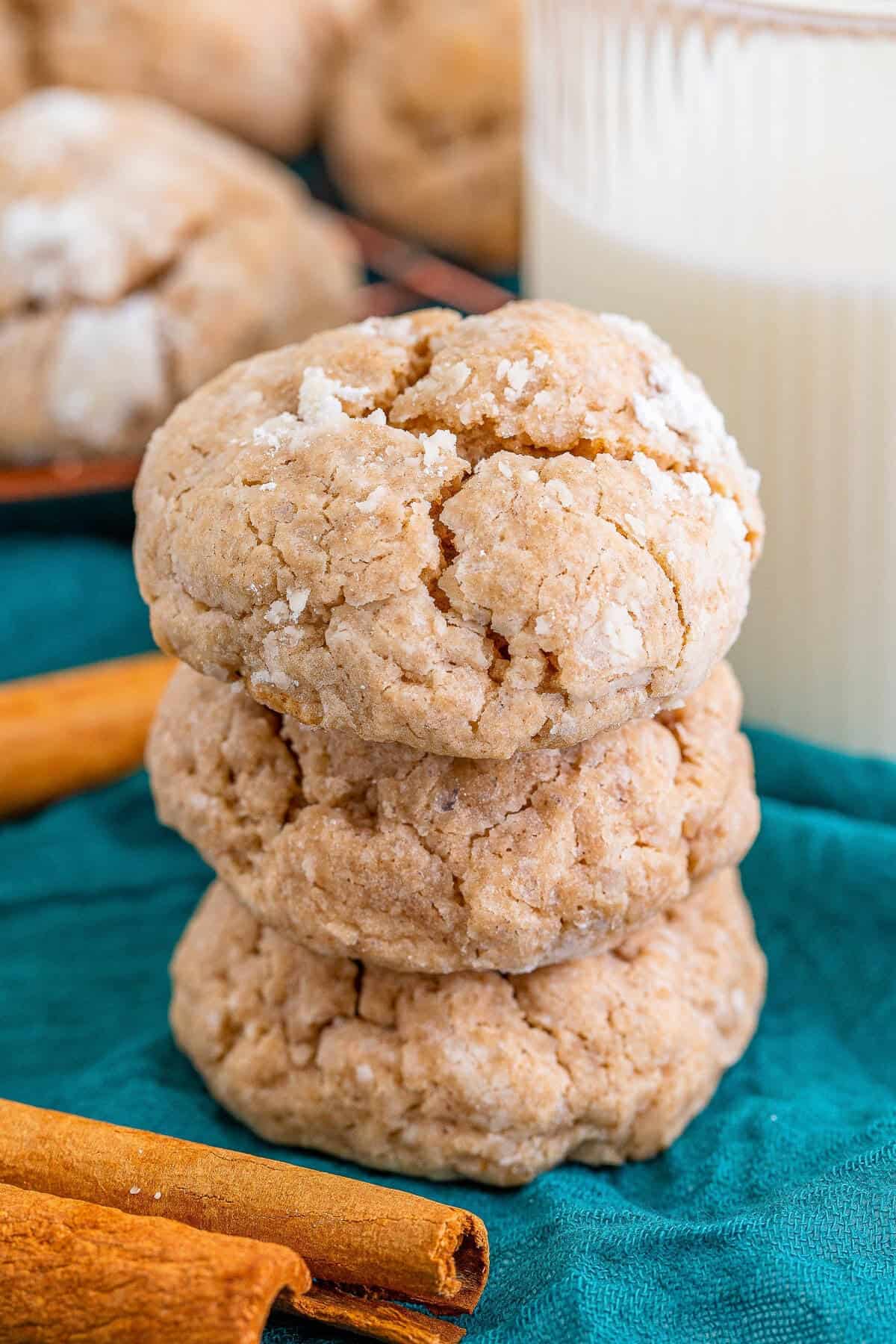 A stack of spiced gooey butter cookies coated in powdered sugar with cinnamon sticks and a glass of milk.