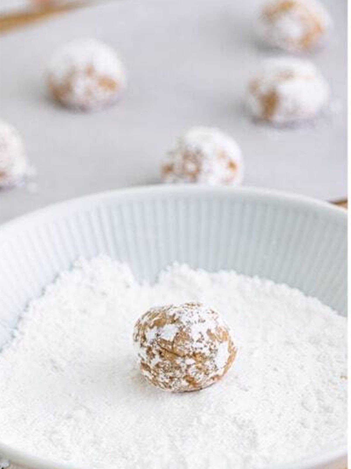 A ball of gooey butter cookie dough being rolled in a bowl of powdered sugar.