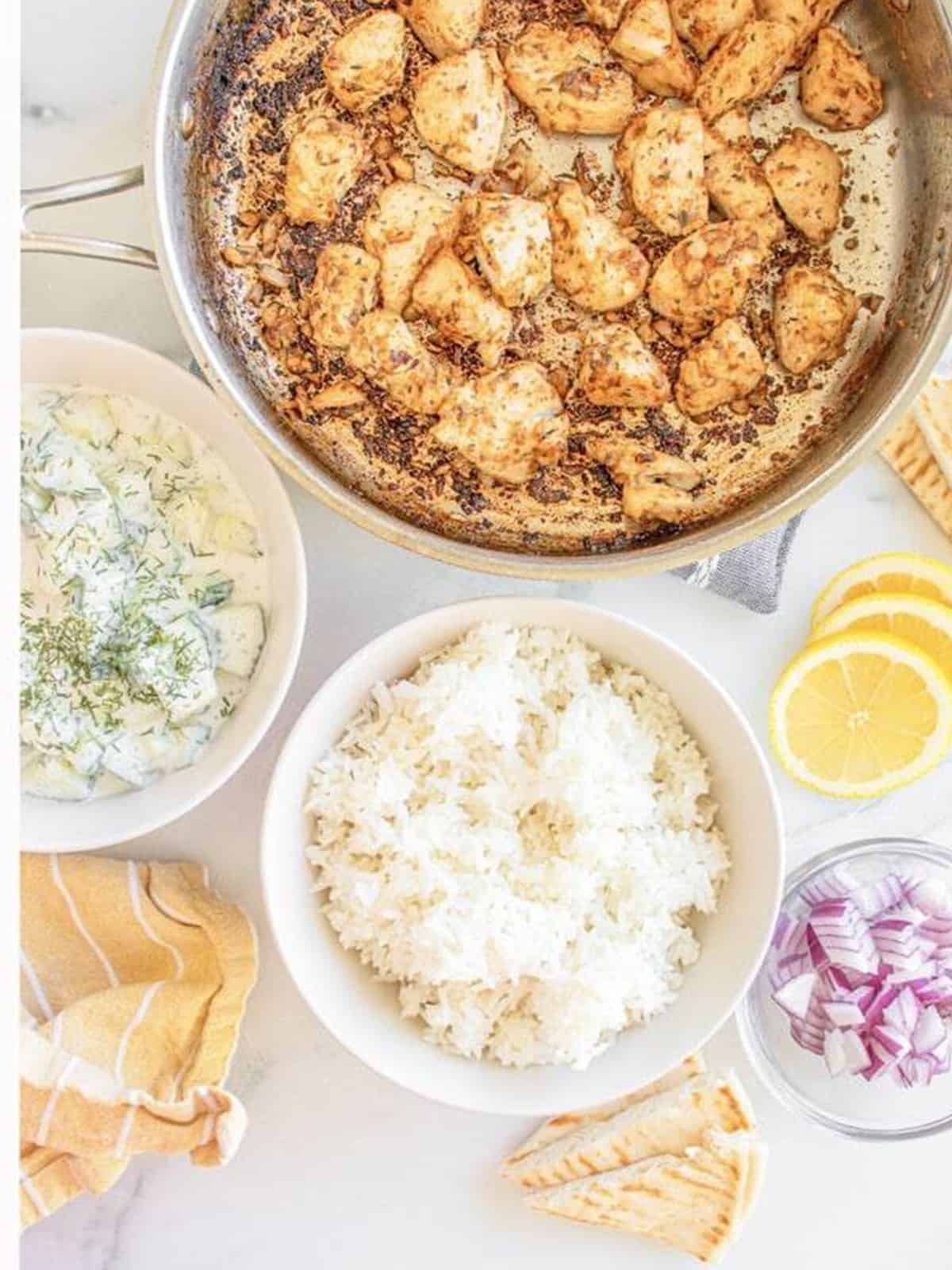 Golden seared Greek chicken in a skillet next to bowls of tzatziki, rice, red onion, and pita bread.