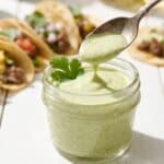 Creamy Green Taco Sauce in a glass jar with a spoon drizzle and street tacos in the background.
