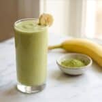 A tall glass of creamy green tea smoothie garnished with a banana slice next to a ripe banana and a bowl of matcha powder.