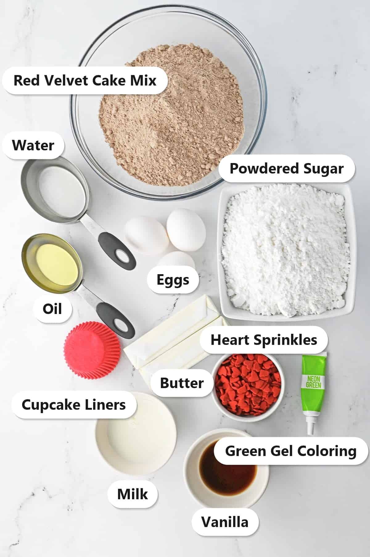 Ingredients to make grinch cupcakes labeled on a marble surface: red velvet cake mix, water, oil, eggs, powdered sugar, butter, cupcake liners, heart sprinkles, green gel coloring, milk, and vanilla.
