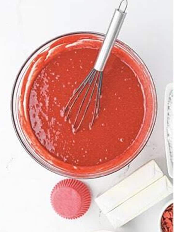 Red velvet cake batter whisked together in a glass bowl with a red cupcake liner nearby.