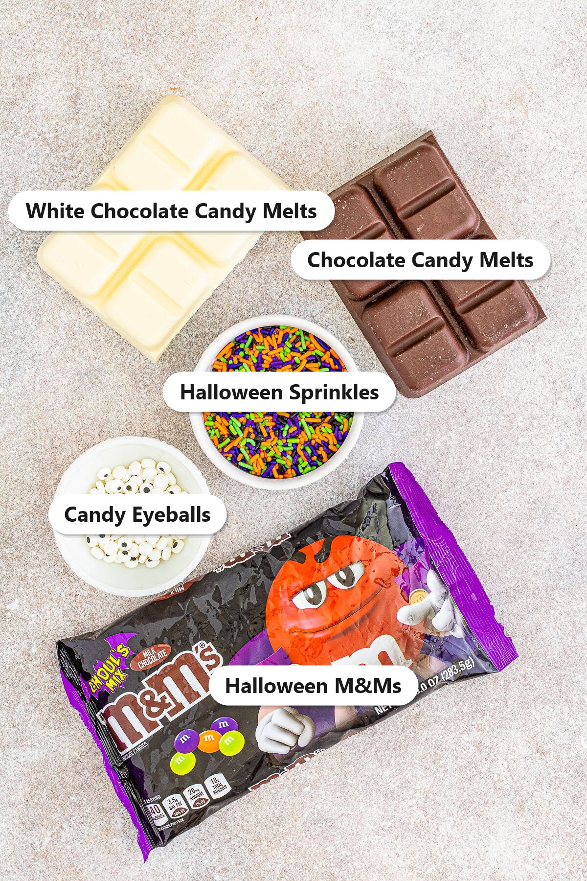 Labeled ingredients for halloween bark including white chocolate candy melts, chocolate candy melts, Halloween M&Ms, Halloween sprinkles, and candy eyeballs.