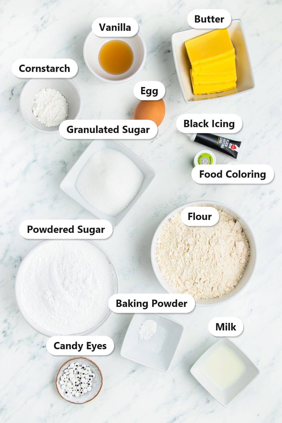 Labeled ingredients for Halloween sugar cookies including butter, granulated sugar, egg, vanilla, flour, cornstarch, baking powder, powdered sugar, milk, food coloring, black icing, and candy eyes.