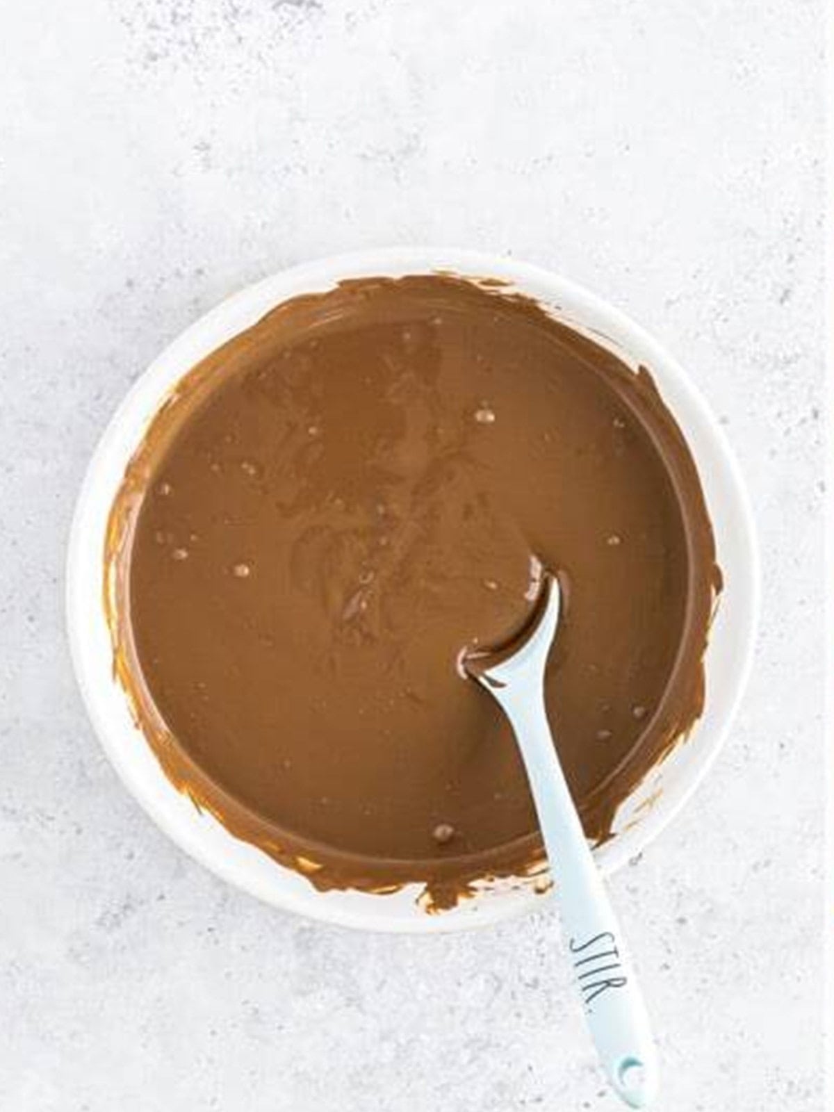 Melted chocolate stirred smooth in a white bowl with a spoon for Hot Cocoa Bombs.