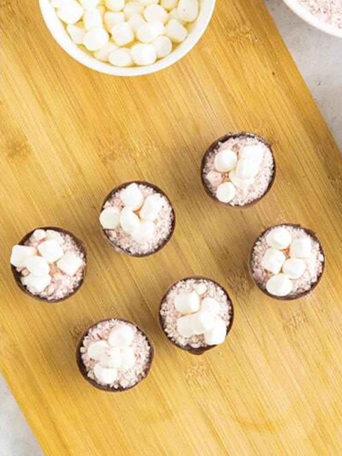 Chocolate shells filled with hot cocoa mix, crushed peppermint, and mini marshmallows on a cutting board.