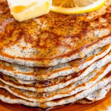 Lemon glaze being poured over a stack of lemon poppy seed pancakes with fresh lemons and berries nearby.