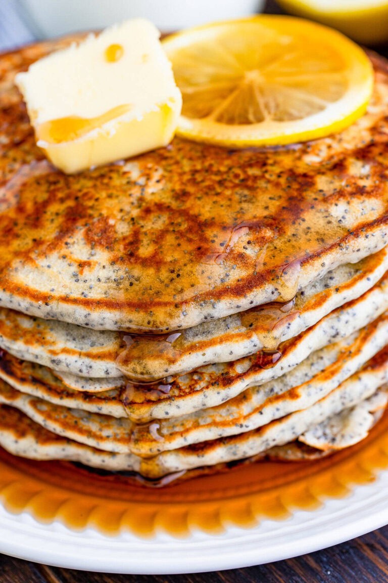 Lemon glaze being poured over a stack of lemon poppy seed pancakes with fresh lemons and berries nearby.