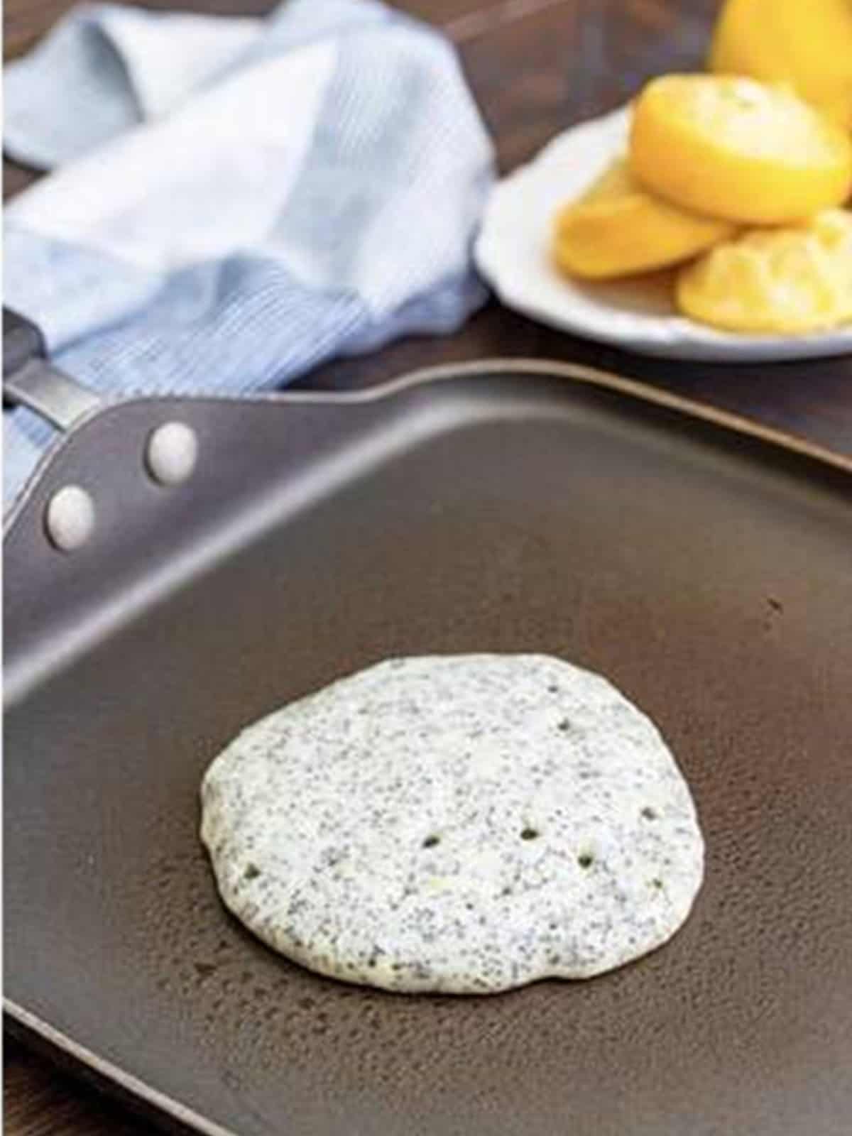 A lemon poppy seed pancake cooking on the griddle with bubbles forming in the center.
