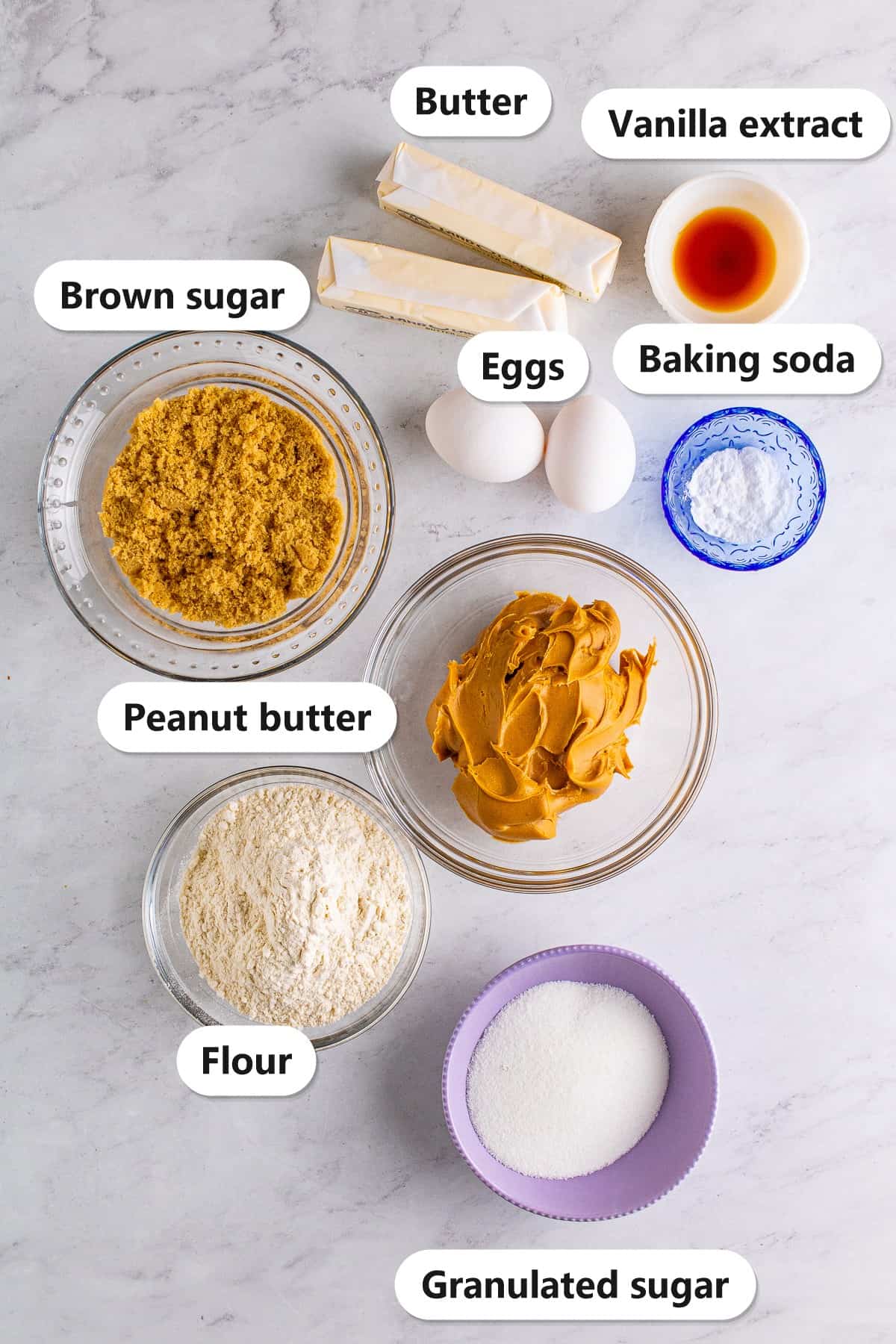 Labeled ingredients for crispy peanut butter cookies including peanut butter, butter, brown sugar, granulated sugar, eggs, vanilla extract, flour, and baking soda on a marble surface.