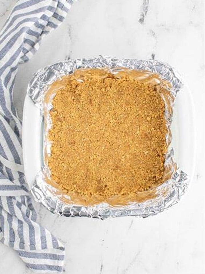 Graham cracker crust pressed into a foil lined square baking dish for maple cheesecake bars.