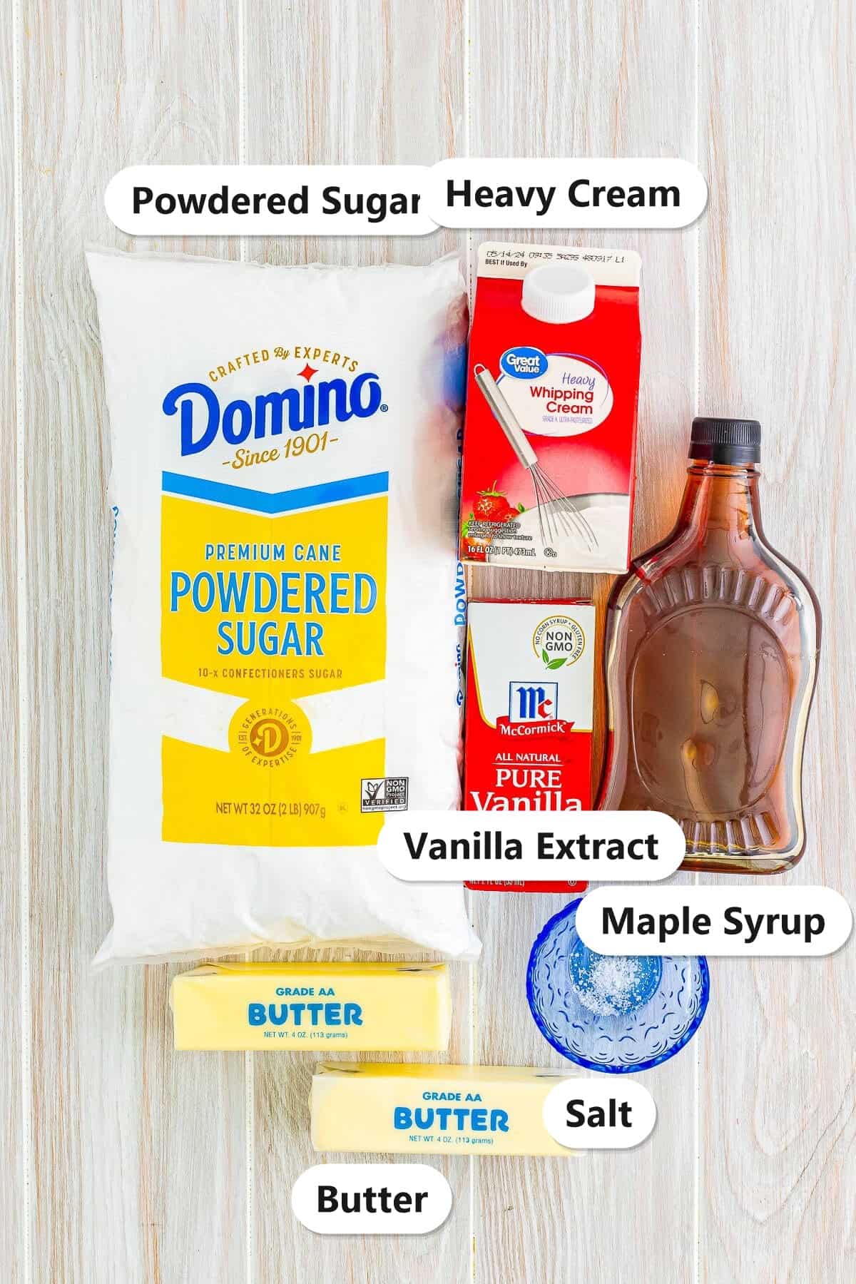 Ingredients to make maple frosting labeled on a white wood surface: powdered sugar, heavy cream, maple syrup, vanilla extract, salt, and butter.