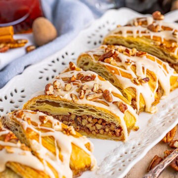 A Maple Pecan Pastry Braid on a white platter drizzled with maple glaze and topped with chopped pecans.