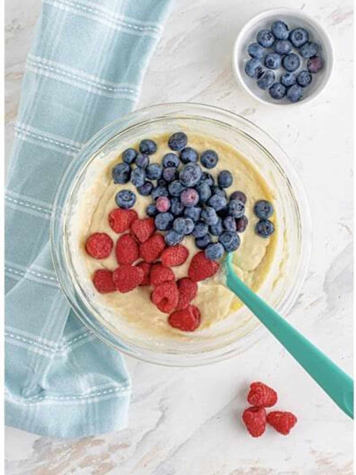 Fresh blueberries and raspberries being folded into pancake batter in a glass bowl.