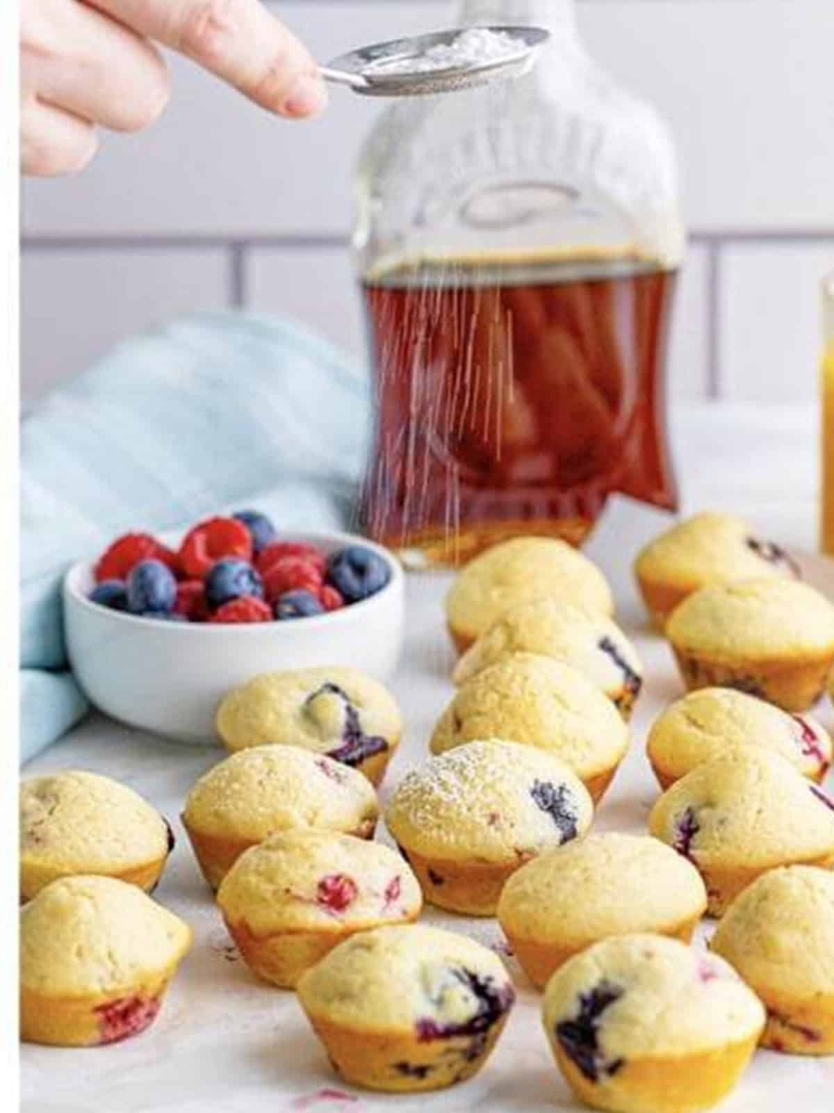 Baked mini berry pancake bites being dusted with powdered sugar next to maple syrup.