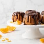 Mini Bundt Cakes with glossy chocolate glaze on a white cake stand with fresh orange slices.