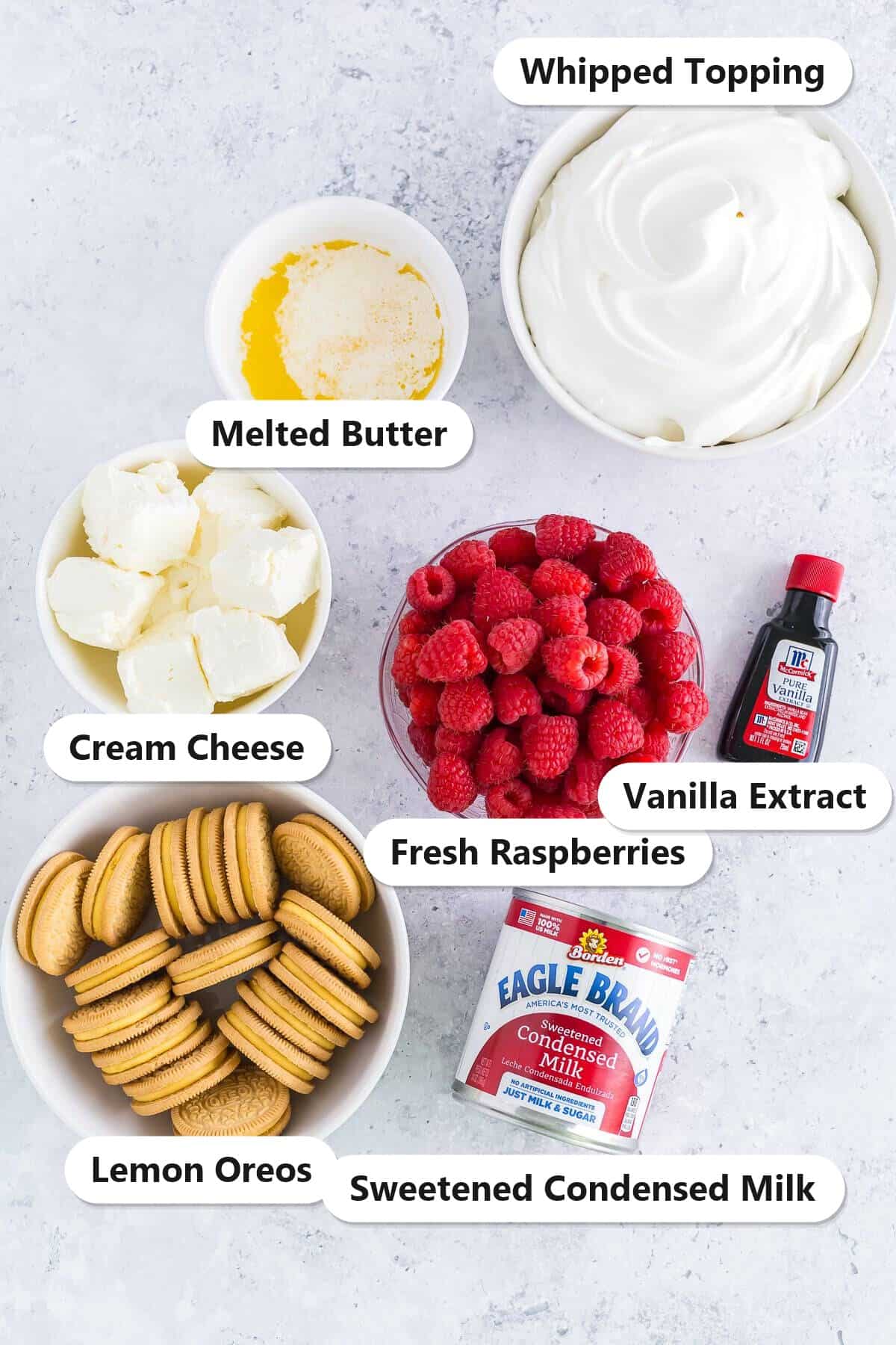 Labeled ingredients for mini lemon cheesecakes including cream cheese, whipped topping, fresh raspberries, lemon Oreos, sweetened condensed milk, melted butter, and vanilla extract.