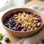 A bowl of no bake blueberry plum crumble with a golden oat and almond crumble topping.