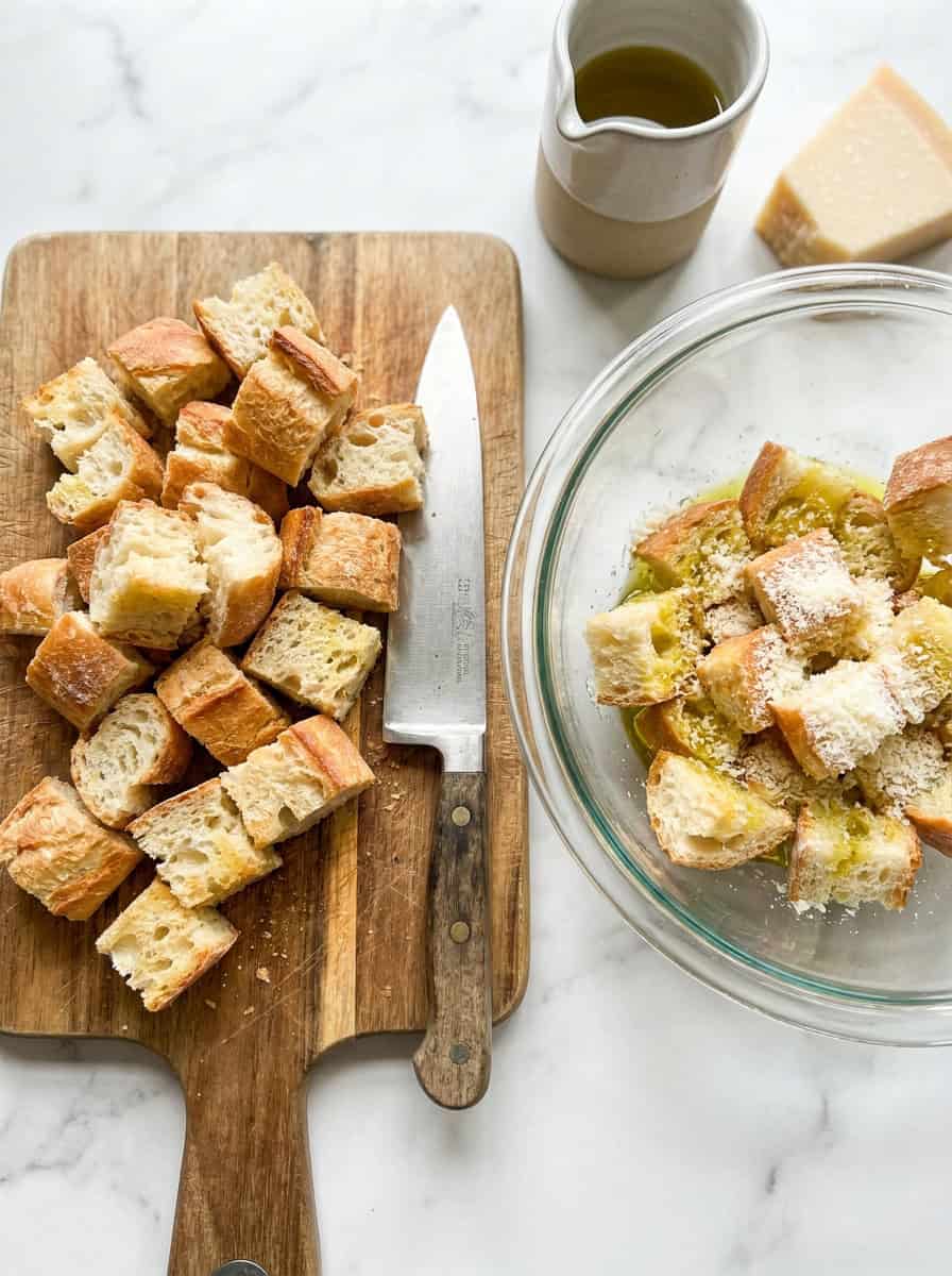 Cubes of french bread on a cutting board tossed with olive oil and parmesan for Panzanella croutons.