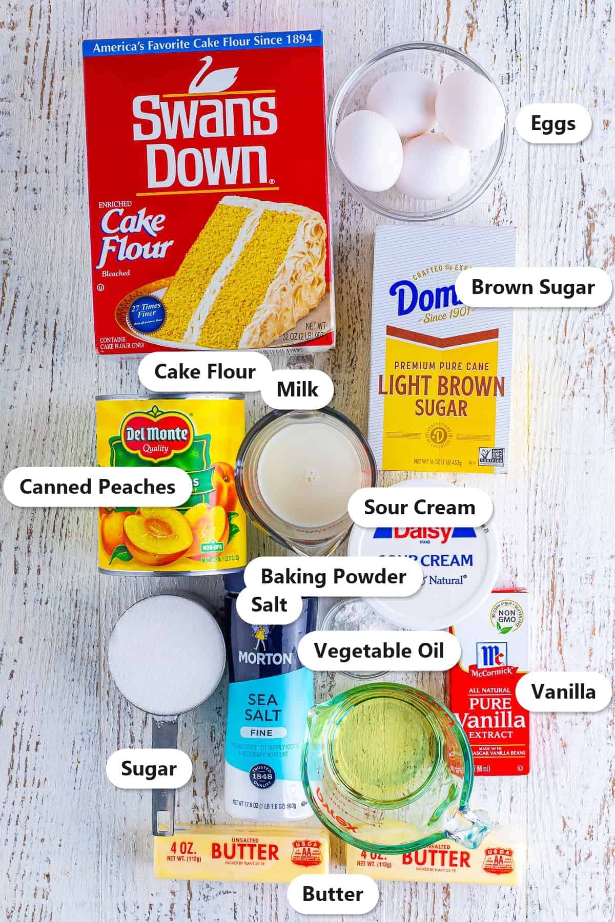 Labeled ingredients to make peach upside down cake including cake flour, canned peaches, brown sugar, eggs, butter, sour cream, and vanilla