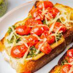 Pesto Toast topped with blistered cherry tomatoes and shredded parmesan on a white plate.