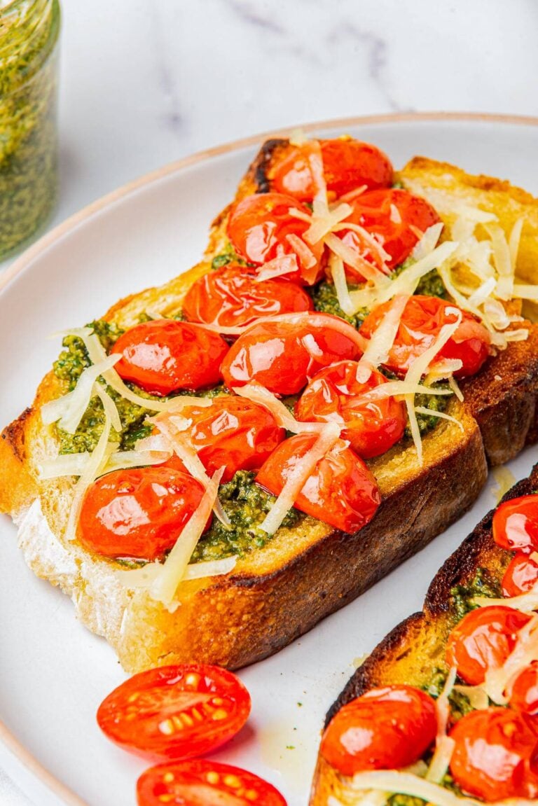 Pesto Toast topped with blistered cherry tomatoes and shredded parmesan on a white plate.