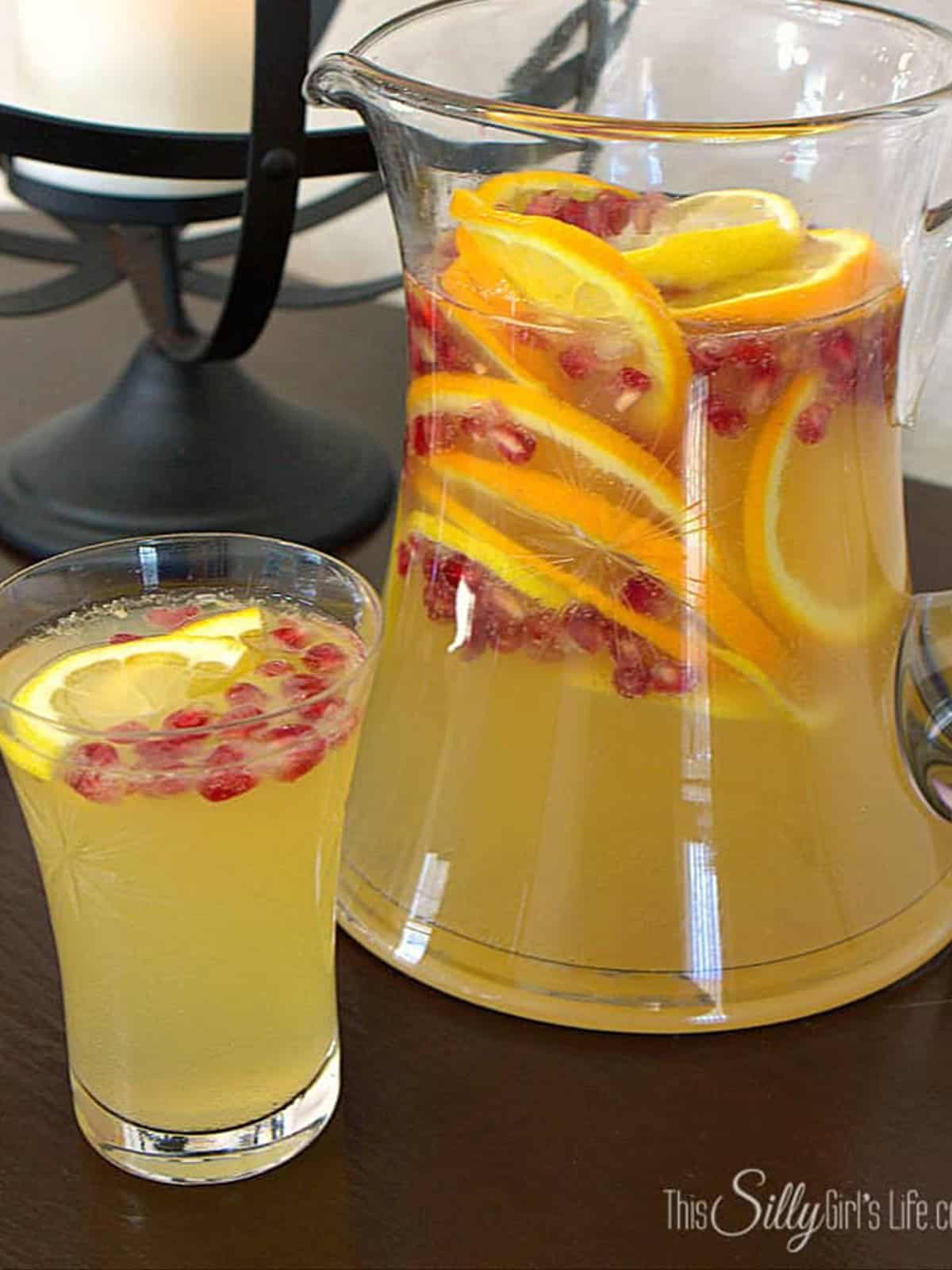 A glass pitcher of pomegranate punch filled with orange slices, lemon slices, and pomegranate seeds next to a filled punch glass.